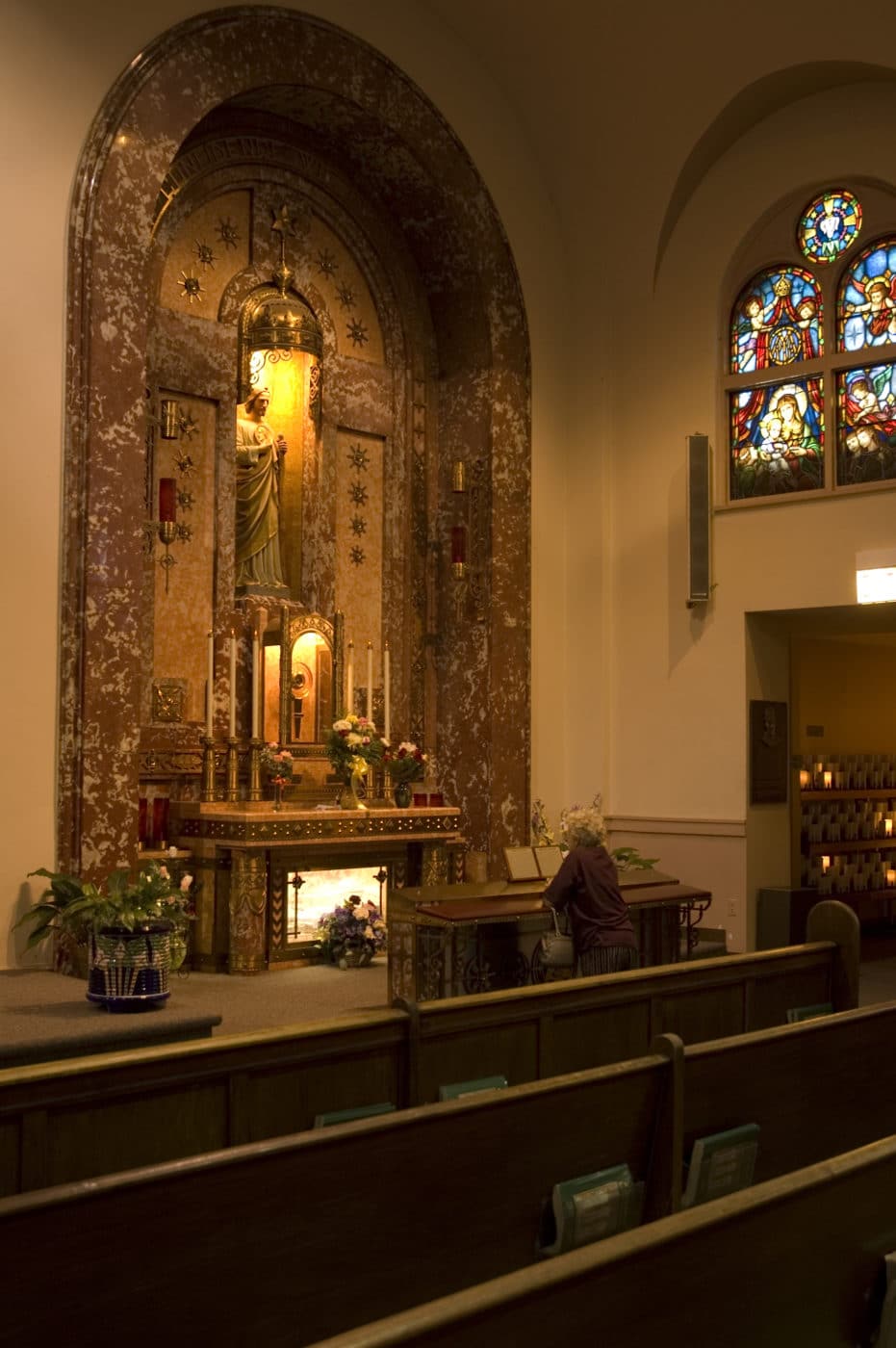 An ornate shrine featuring a statue of St. Jude in a niche, surrounded by intricate designs and patterns with ornamental accents. Several candles are placed at the base, along with red glass candle holders on the sides. The statue holds a staff and has a halo around its head.
