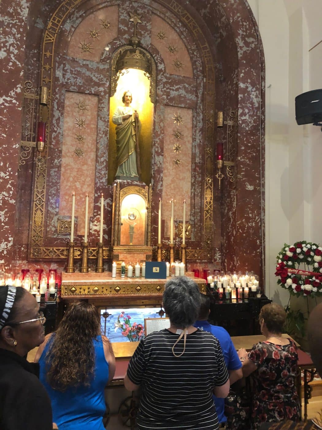A group of people stand and pray in front of an ornate altar featuring a statue of St. Jude, surrounded by lit candles and floral arrangements. The background is adorned with intricate designs and patterns.