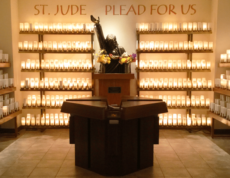 A statue of St. Jude with one arm raised and the other hand holding an image of Jesus to his chest, surrounded by rows of lit candles on shelves in the background. Text on the back wall reads 'ST. JUDE PLEAD FOR US'.