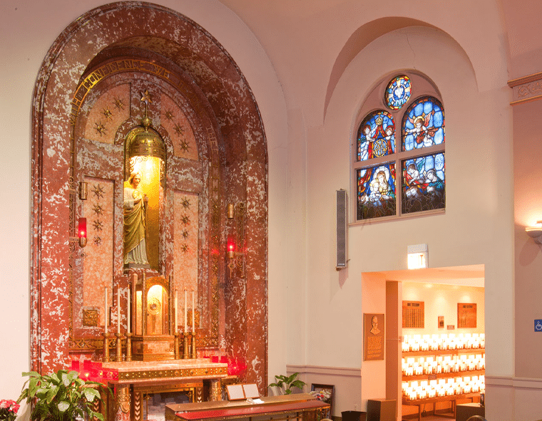 Interior view of the church sanctuary at The National Shrine of St. Jude that features an ornate altar with a statue of St. Jude, surrounded by candles and decorative elements. A doorway to the right reveals additional lit candles, and a stained glass window is positioned above the doorway.