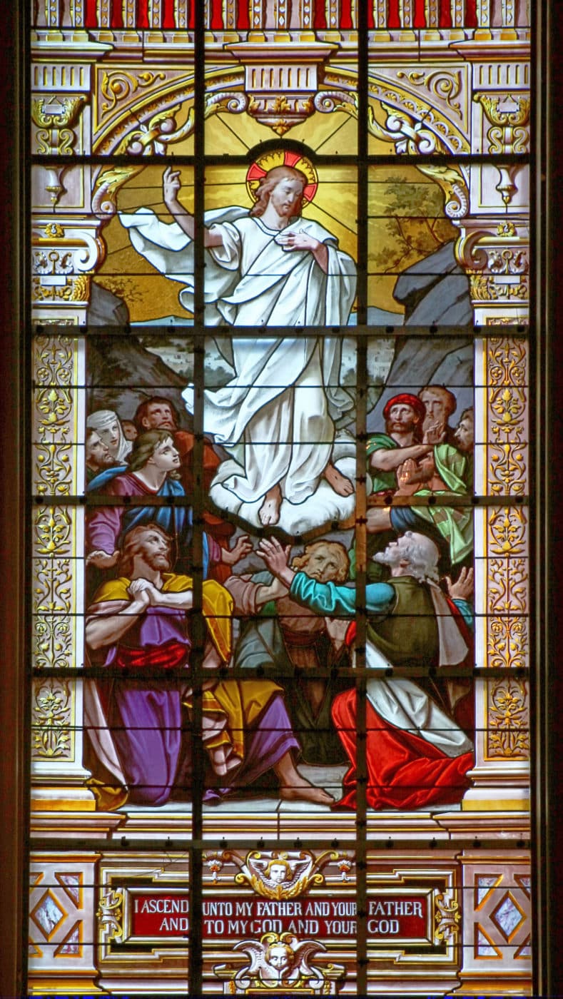 A stained glass window depicting a religious scene with a central figure ascending while surrounded by several other figures looking up in reverence. Ornate architectural elements frame the scene, and text at the bottom reads: 'I ASCEND UNTO MY FATHER AND YOUR FATHER AND TO MY GOD AND YOUR GOD.'