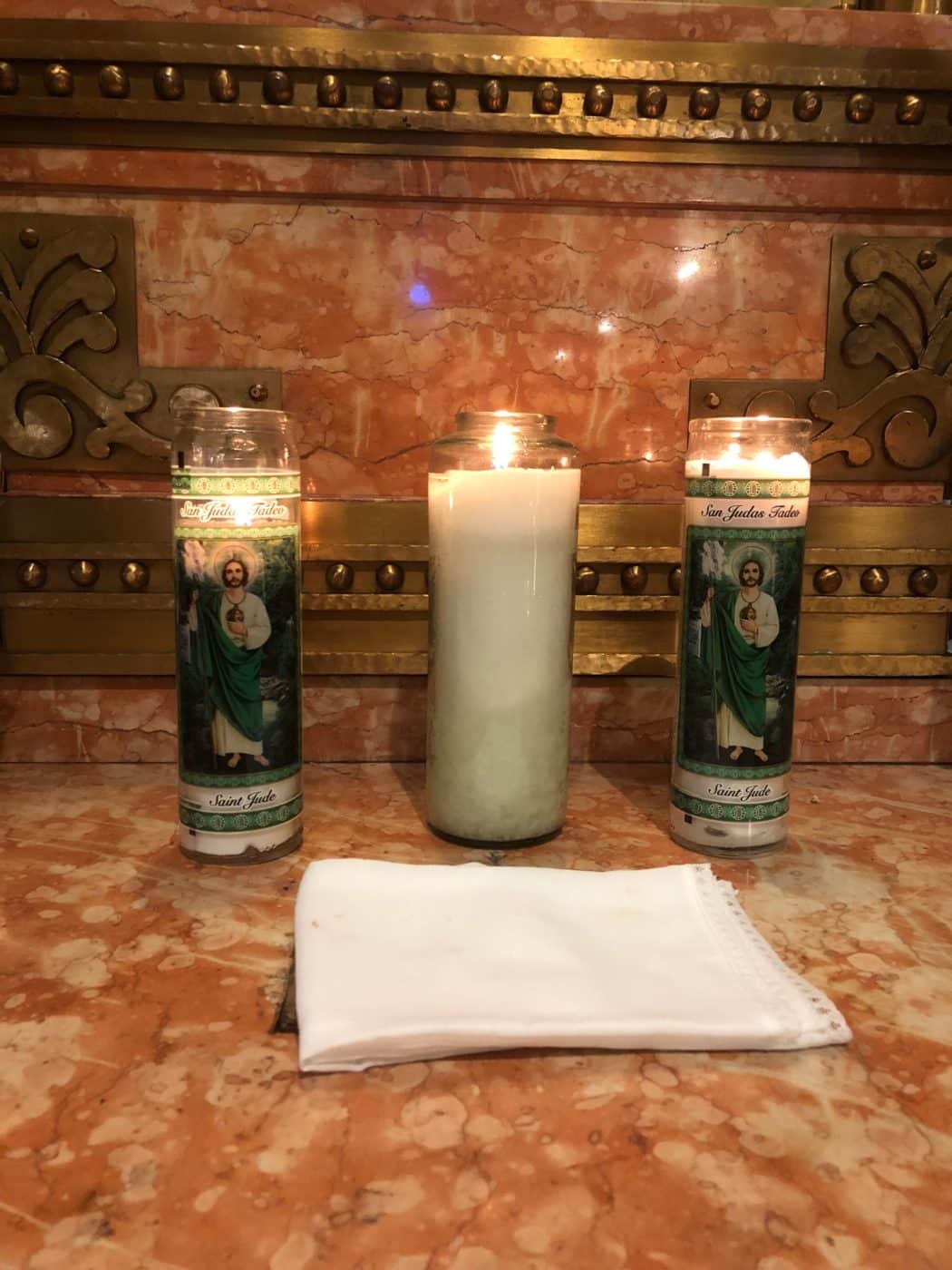 An altar with three lit candles; two candles feature images of Saint Jude and are placed on either side of a plain candle in the center. A folded white cloth lies in front of the candles on a marble surface, with ornate decorative elements in the background.