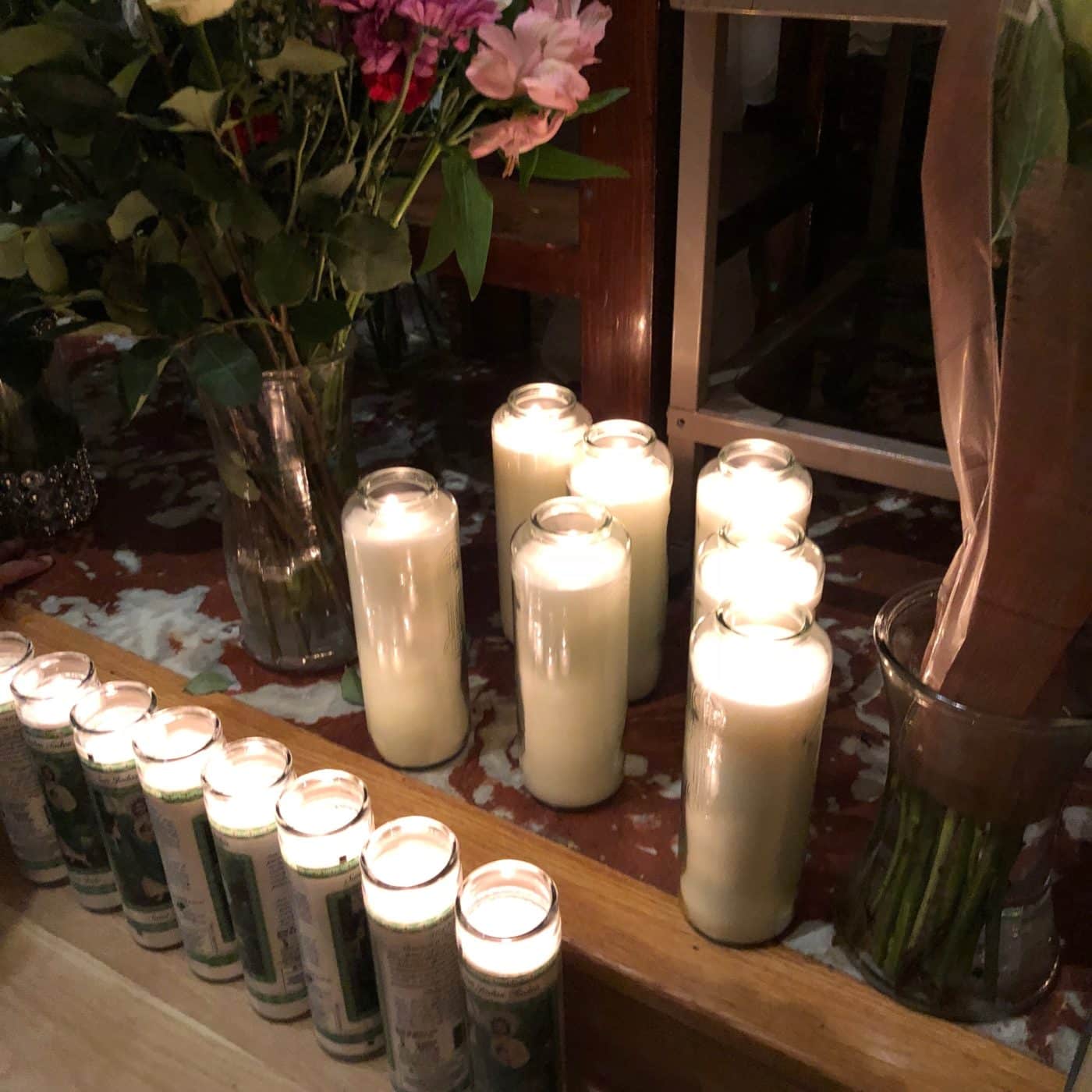 A group of lit prayer candles arranged on an altar, with some in glass holders at the Shrine of St. Jude. Vases with flowers and stems are placed nearby.