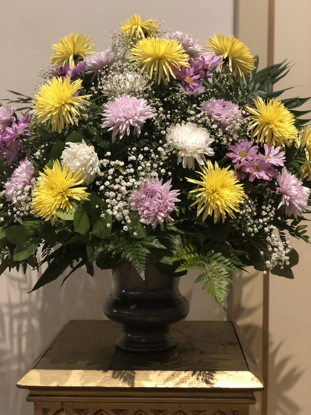 A large floral arrangement in a dark vase on a wooden table. The bouquet includes chrysanthemums and baby's breath, surrounded by green leaves.